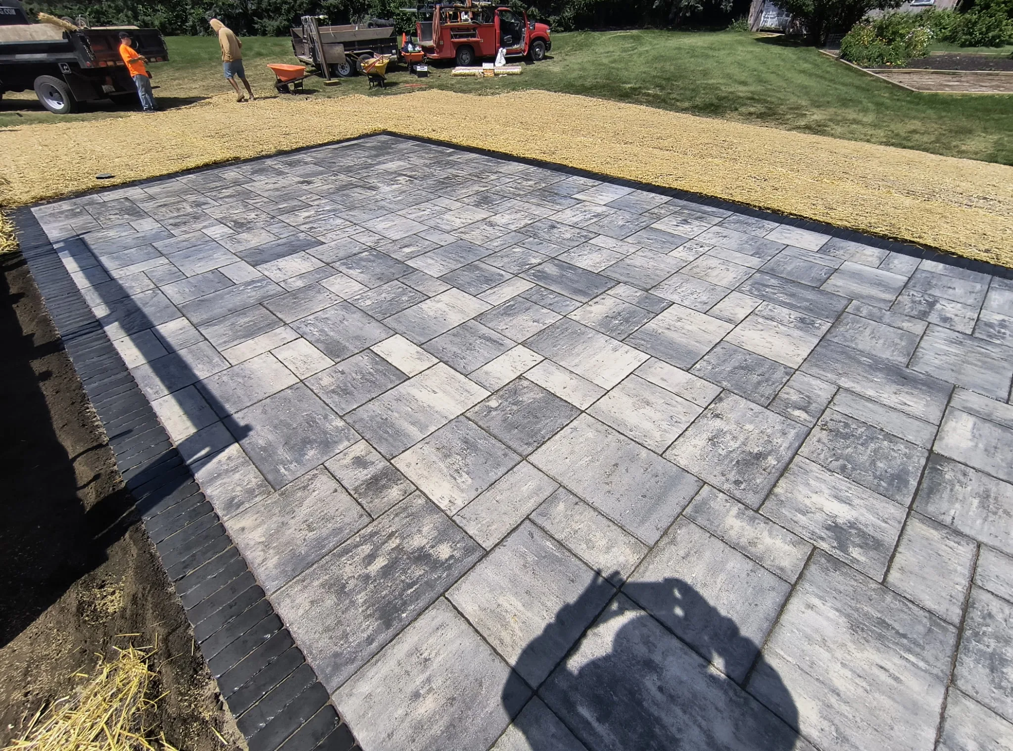 Newly installed gray paver patio with gravel border, crew and equipment working in the background